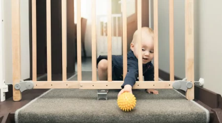 Kind spielt mit einem Ball hinter einem Schutzgitter aus Holz vor einer Treppe
