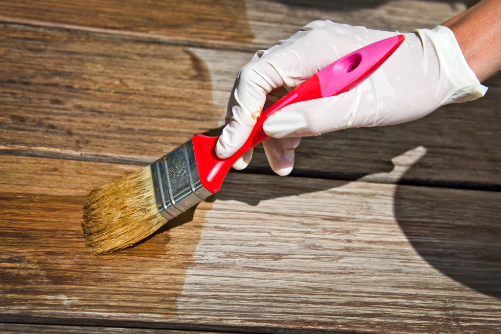 Hand mit Latexhandschuh und rotem Pinsel streicht mit einer Lasur über Bambusdielen einer Terrasse