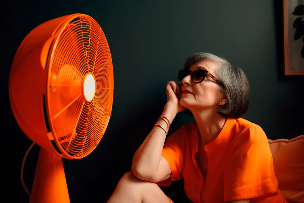 Frau mit orangenem Oberteil sitzt vor einem orangen Ventilator und lässt sich abkühlen