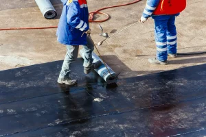 Zwei Arbeiter bringen mit einem Gasbrenner eine Bitumen-Schweißbahn auf einem Flachdach auf – typische Abdichtungsmaßnahme im Dachdeckerhandwerk.