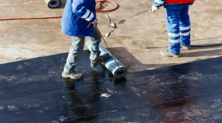 Zwei Arbeiter bringen mit einem Gasbrenner eine Bitumen-Schweißbahn auf einem Flachdach auf – typische Abdichtungsmaßnahme im Dachdeckerhandwerk.