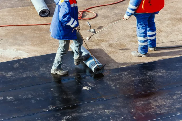 Zwei Arbeiter bringen mit einem Gasbrenner eine Bitumen-Schweißbahn auf einem Flachdach auf – typische Abdichtungsmaßnahme im Dachdeckerhandwerk.