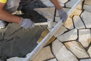 Fliesenleger verlegt Natursteinplatten im Garten auf Betonfundament mit Gummihammer und Wasserwaage