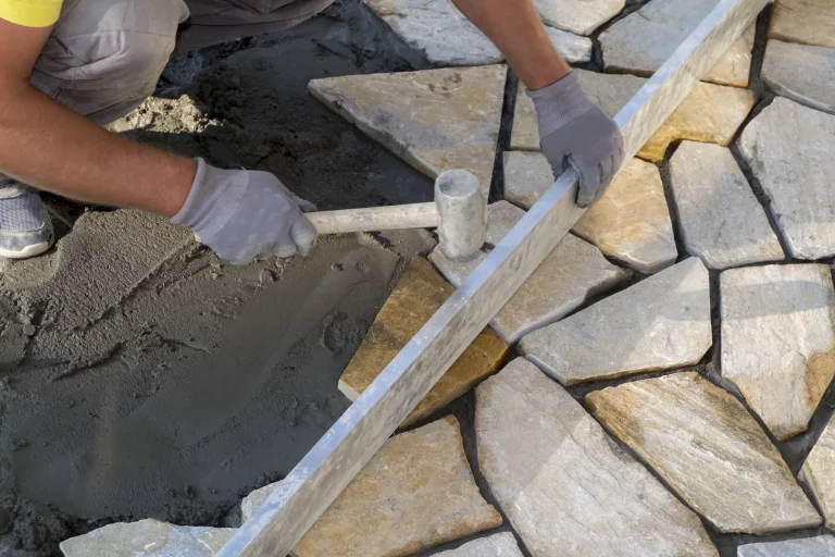 Fliesenleger verlegt Natursteinplatten im Garten auf Betonfundament mit Gummihammer und Wasserwaage
