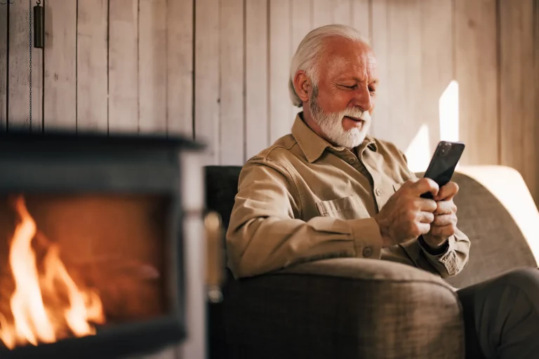 Älterer Mann sitzt mit einem Smartphone vor einem brennenden Kaminofen