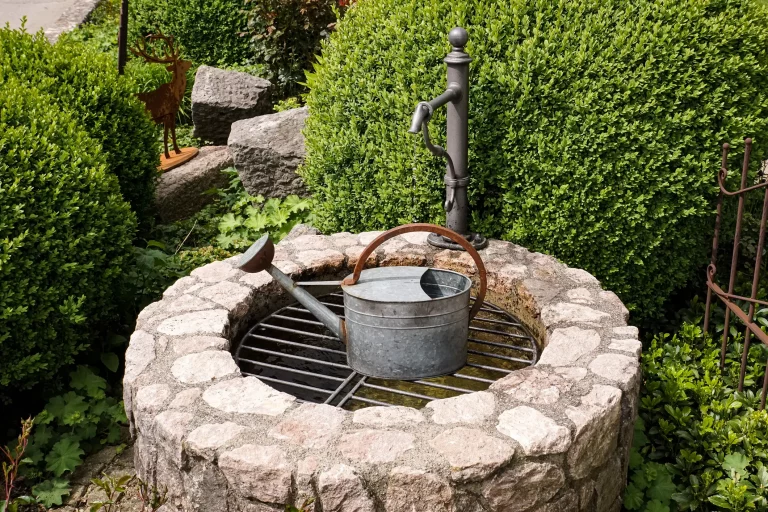 Alter Brunnen in einem Garten mit einem Hebel zum Pumpfen von Wasser und einer alten Gieskanne