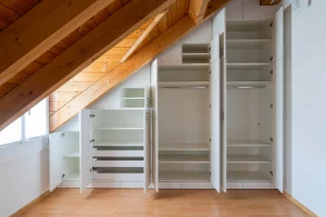 Eine Innenansicht eines maßgefertigten Einbauschranks mit offenen Türen, die in ein Schlafzimmer mit Dachschräge eingebaut sind