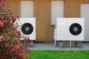 Zwei weiße Außengeräte einer Luftwärmepumpe auf Betonsockeln vor einem modernen Holzhaus mit geschlossenen Rollläden und blühendem Busch im Vordergrund.