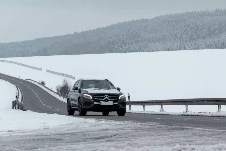 Schwarzer Mercedes-SUV fährt auf einer schneebedeckten Landstraße mit bewaldeter Winterlandschaft im Hintergrund, Fokus auf Fahrsicherheit bei Eis und Schnee.