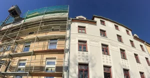 Zwei nebeneinander stehende Gebäude mit vorher und nachher Effekt einer Fassadensanierung. Das Haus links mit Gerüst vor der Sanierung der Fassade. Das Haus rechts mit fertig sanierter Fassade.