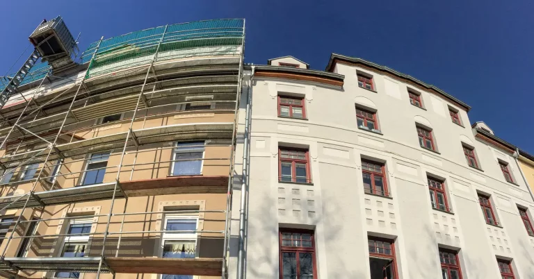 Zwei nebeneinander stehende Gebäude mit vorher und nachher Effekt einer Fassadensanierung. Das Haus links mit Gerüst vor der Sanierung der Fassade. Das Haus rechts mit fertig sanierter Fassade.