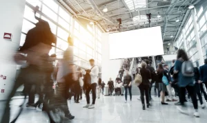 Besucher einer Messe fahren Rolltreppen hoch und runter. An der Decke hängen Elemente des Messebaus an der eine Leinwand befästigt wurde, die über der Rolltreppe hängt.