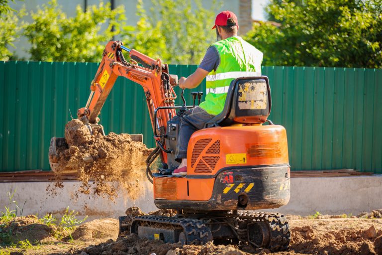 Baggerfahrer auf einem orangenen Minibagger in einem Garten mit Erde in der Schaufel