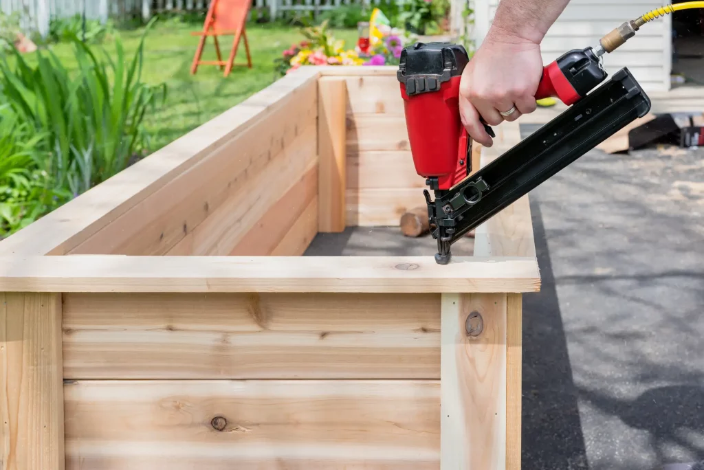 Ein Mann baut ein Hochbeet aus Holz und befestigt mit einer Nagelpistole Holzbretter