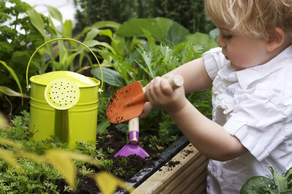 Kleinkind steht mit Gieskanne und einer orangenen kleinen Gartenschaufel an einem Hochbeet