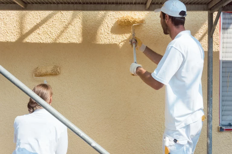 Ein Maler und eine Malerin in weißer Malerkleidung tragen mit Malerrollen Rollputz an einer Außenfassade auf