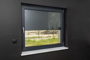 Modernes Fenster mit halb heruntergelassenem elektrischen Rollladen in einem Raum mit dunkler Wandfarbe. Links neben dem Fenster befindet sich ein schwarzer Schalter zur Bedienung der Rollläden. Draußen ist ein grüner Rasen und ein Baugrundstück zu sehen. Das Bild illustriert die Nachrüstung von elektrischen Rollläden zur Verbesserung der Fensterisolierung und des Komforts.