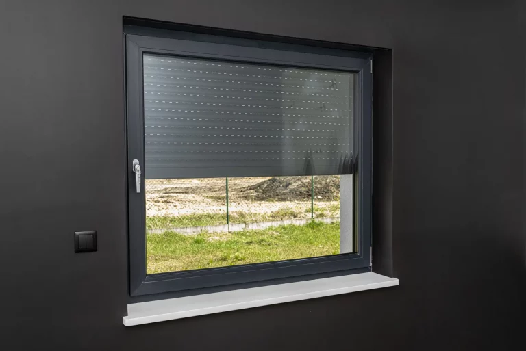 Modernes Fenster mit halb heruntergelassenem elektrischen Rollladen in einem Raum mit dunkler Wandfarbe. Links neben dem Fenster befindet sich ein schwarzer Schalter zur Bedienung der Rollläden. Draußen ist ein grüner Rasen und ein Baugrundstück zu sehen. Das Bild illustriert die Nachrüstung von elektrischen Rollläden zur Verbesserung der Fensterisolierung und des Komforts.