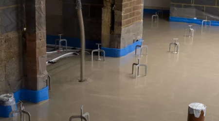 Ein frischer Estrichboden in einem Rohbau trocknet gleichmäßig. Mehrere Installationspunkte und Rohre ragen aus dem noch feuchten Estrich heraus. Blaue Schutzfolien an den Wänden und um die Pfeiler schützen die Umgebung während des Trocknungsprozesses. Das Bild verdeutlicht die Phase der Trocknung des Estrichs, die wichtig ist, um eine stabile und tragfähige Bodenfläche zu gewährleisten.