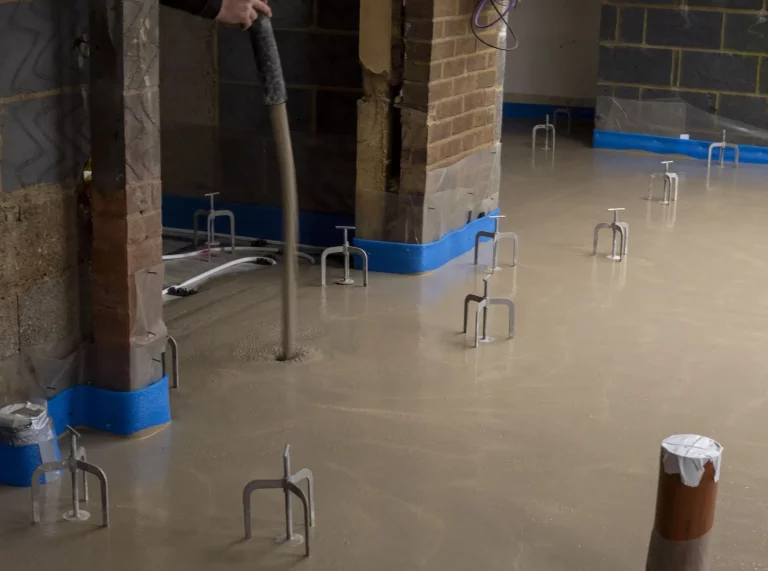 Ein frischer Estrichboden in einem Rohbau trocknet gleichmäßig. Mehrere Installationspunkte und Rohre ragen aus dem noch feuchten Estrich heraus. Blaue Schutzfolien an den Wänden und um die Pfeiler schützen die Umgebung während des Trocknungsprozesses. Das Bild verdeutlicht die Phase der Trocknung des Estrichs, die wichtig ist, um eine stabile und tragfähige Bodenfläche zu gewährleisten.