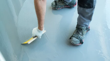 Ein Estrichleger glättet eine frische Estrichschicht mit einer Kelle. Er trägt Arbeitshandschuhe und schmutzige Arbeitsschuhe mit Überziehern, die auf dem glatten Estrichboden stehen. Die Oberfläche des Estrichs zeigt noch Spuren der Verteilung, was den Verlegeprozess verdeutlicht. Das Bild veranschaulicht die handwerkliche Tätigkeit und die sorgfältige Ausführung bei der Verlegung von Estrich.
