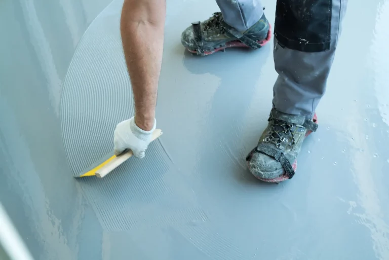 Ein Estrichleger glättet eine frische Estrichschicht mit einer Kelle. Er trägt Arbeitshandschuhe und schmutzige Arbeitsschuhe mit Überziehern, die auf dem glatten Estrichboden stehen. Die Oberfläche des Estrichs zeigt noch Spuren der Verteilung, was den Verlegeprozess verdeutlicht. Das Bild veranschaulicht die handwerkliche Tätigkeit und die sorgfältige Ausführung bei der Verlegung von Estrich.