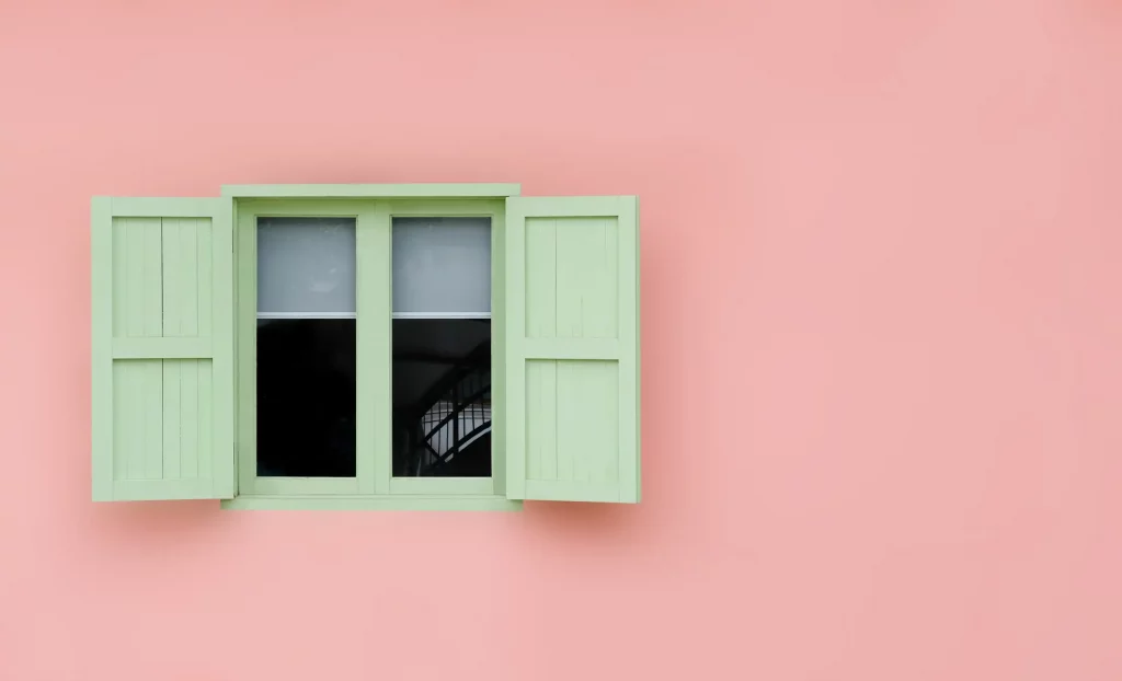 Hellgrüne Fensterrahmen aus Holz mit hellgrünen Fensterläden auf einer rosa farbigen Fassade