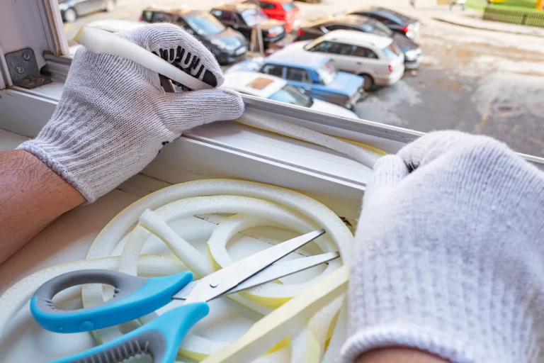 Ein Fensterbauer mit weißen Handschuhen, die Schaumstoffdichtungen an einem Fensterrahmen anbringt. Im Vordergrund sind eine Schere und zusätzliche Schaumstoffdichtung für die Abdichtung von Fenstern zu sehen. Im Hintergrund sind unscharf parkende Autos sichtbar.