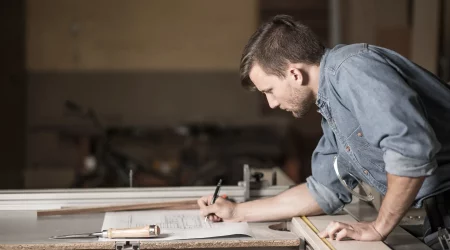Ein Schreiner in einer Werkstatt zeichnet präzise Pläne für eine maßgefertigte Küche. Er trägt ein Jeanshemd und konzentriert sich auf die Skizze, die auf einer Werkbank liegt. Neben ihm liegen Werkzeuge wie ein Zollstock und ein Stechbeitel. Das Bild zeigt den Planungs- und Designprozess für individuelle Küchenanfertigungen vom Schreiner.
