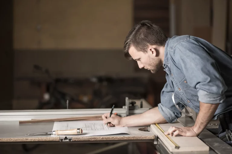 Ein Schreiner in einer Werkstatt zeichnet präzise Pläne für eine maßgefertigte Küche. Er trägt ein Jeanshemd und konzentriert sich auf die Skizze, die auf einer Werkbank liegt. Neben ihm liegen Werkzeuge wie ein Zollstock und ein Stechbeitel. Das Bild zeigt den Planungs- und Designprozess für individuelle Küchenanfertigungen vom Schreiner.