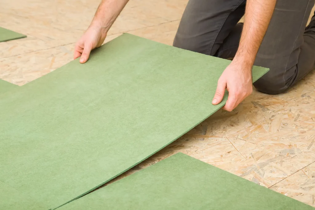 Ein Handwerker, kniet auf einem Boden und verlegt eine grüne Naturholzfaser-Dämmplatte als Trittschalldämmung für Laminat auf einen OSB-Holzboden. Die Hände der Person halten die Dämmplatte an den Ecken, während sie diese in Position bringt. Die Umgebung ist gut beleuchtet und zeigt keine weiteren Gegenstände. Der Fokus liegt auf der Tätigkeit des Verlegens der Platten.