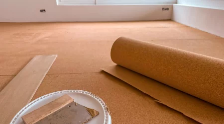Verlegung einer Trittschalldämmung aus Kork unter Laminatboden in einem Raum mit großen Fenstern. Eine Rolle der Dämmung liegt ausgerollt auf dem Boden, daneben ein Eimer mit Werkzeug zur Verarbeitung.