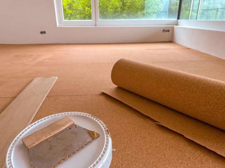 Verlegung einer Trittschalldämmung aus Kork unter Laminatboden in einem Raum mit großen Fenstern. Eine Rolle der Dämmung liegt ausgerollt auf dem Boden, daneben ein Eimer mit Werkzeug zur Verarbeitung.