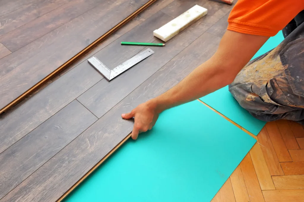 Ein Schreiner, der Laminat auf einer Trittschalldämmung verlegt. Die Trittschalldämmung besteht aus einer türkisfarbenen Schaumstoffunterlage. Der Schreiner trägt eine orangefarbene Arbeitskleidung und verwendet seine Hände, um die Laminatdielen passgenau zu verlegen. Neben ihm liegen ein Bleistift, ein Winkelmaß und eine weiße Kunststoffleiste als Werkzeuge bereit. Der Hintergrund besteht aus bereits verlegtem Laminat und einem Abschnitt eines Parkettbodens.
