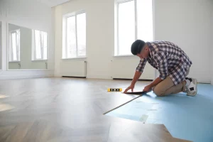 Ein Mann in kariertem Hemd und hellen Hosen kniet in einem hellen Raum und verlegt Holzboden auf bereits verlegtem Trockenestrich. Der Raum hat große Fenster und weiße Wände, mit einem Heizkörper unter einem der Fenster. Der Mann benutzt eine Wasserwaage, um die Verlegung des Holzbodens zu überprüfen. Der Boden ist teilweise mit Holzdielen bedeckt, während ein Teil noch die blaue Unterlage des Trockenestrichs zeigt.