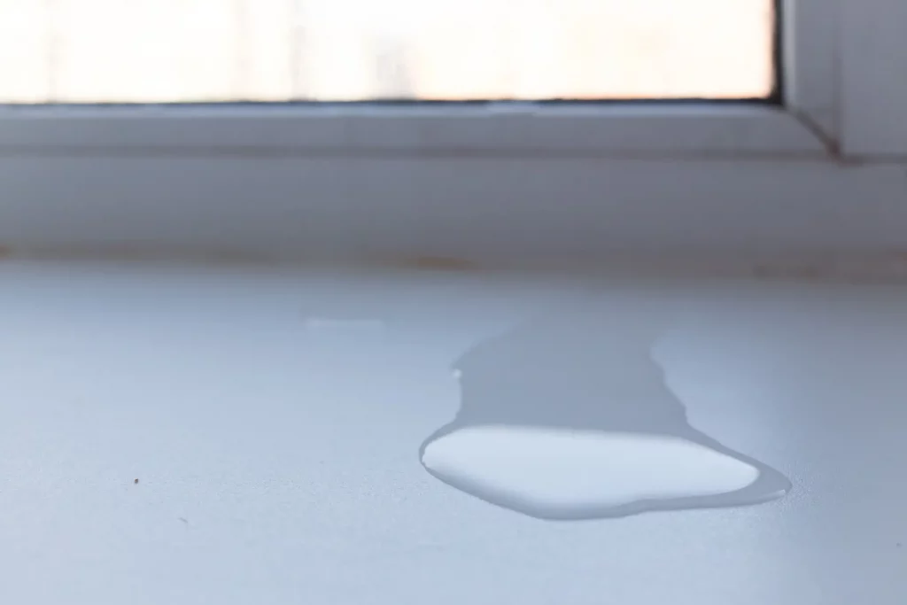 Wasserlache auf einer hellen Fensterbank unter einem geschlossenen, undichten Fenster. Im Hintergrund ist verschwommenes Außenlicht sichtbar.