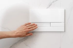 Eine Hand betätigt die Spültaste eines weißen Unterputzspülkastens, der in einer weißen Marmorwand installiert ist. Der moderne Spülkasten ist platzsparend in der Wand integriert und bietet eine elegante Lösung für Badezimmer.