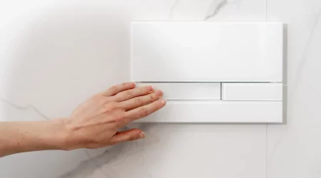 Eine Hand betätigt die Spültaste eines weißen Unterputzspülkastens, der in einer weißen Marmorwand installiert ist. Der moderne Spülkasten ist platzsparend in der Wand integriert und bietet eine elegante Lösung für Badezimmer.
