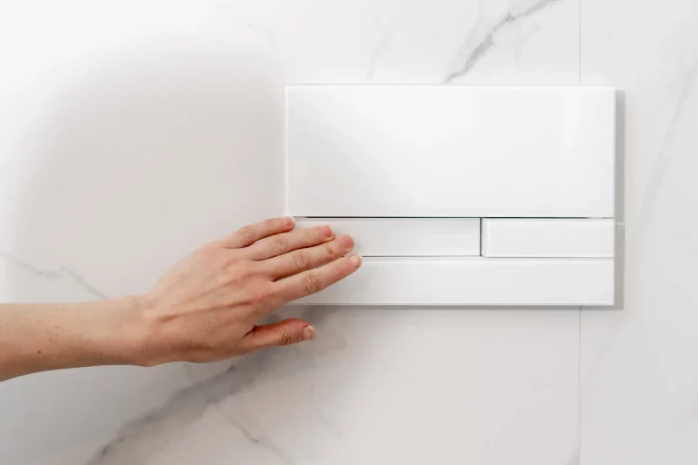 Eine Hand betätigt die Spültaste eines weißen Unterputzspülkastens, der in einer weißen Marmorwand installiert ist. Der moderne Spülkasten ist platzsparend in der Wand integriert und bietet eine elegante Lösung für Badezimmer.