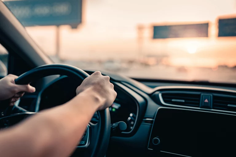 Blick aus dem Fahrzeuginneren auf die Hände einer Person am Lenkrad während der Fahrt auf der Autobahn bei Sonnenuntergang, mit Fokus auf das Armaturenbrett und die Fahrbahn im Hintergrund.