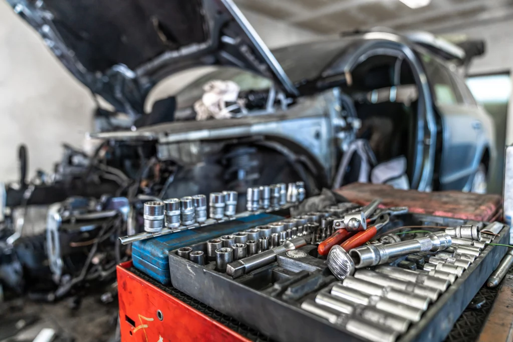 Detailaufnahme eines offenen Werkzeugkastens mit Stecknüssen und Schraubenschlüsseln in einer Autowerkstatt, im Hintergrund ein teilweise zerlegtes Fahrzeug mit geöffneter Motorhaube.