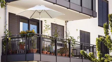 Balkon eines modernen Wohngebäudes mit Außenjalousien, die den Innenraum vor Sonneneinstrahlung schützen. Der Balkon ist mit Pflanzen und einem Sonnenschirm dekoriert, was eine gemütliche Atmosphäre schafft. Die schwarzen Aluminiumjalousien sind teilweise geschlossen, um Privatsphäre zu bieten.