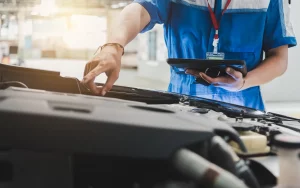 Ein KFZ-Mechaniker in blauer Arbeitskleidung überprüft den Motorraum eines Autos in einer Werkstatt. In der einen Hand hält er ein Tablet zur digitalen Dokumentation und Bewertung, während seine andere Hand im Motorraum arbeitet. Die Szene ist gut beleuchtet und zeigt die moderne Herangehensweise an die Autobewertung und Wartung, bei der digitale Werkzeuge zum Einsatz kommen, um den Zustand des Fahrzeugs zu erfassen und zu bewerten.