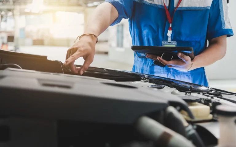 Ein KFZ-Mechaniker in blauer Arbeitskleidung überprüft den Motorraum eines Autos in einer Werkstatt. In der einen Hand hält er ein Tablet zur digitalen Dokumentation und Bewertung, während seine andere Hand im Motorraum arbeitet. Die Szene ist gut beleuchtet und zeigt die moderne Herangehensweise an die Autobewertung und Wartung, bei der digitale Werkzeuge zum Einsatz kommen, um den Zustand des Fahrzeugs zu erfassen und zu bewerten.