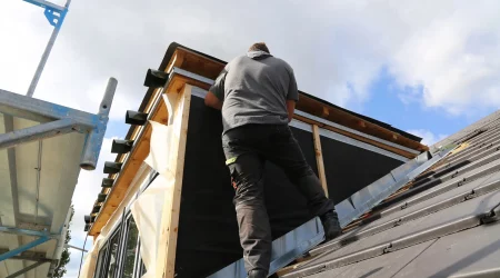 Ein Dachdecker in grauem T-Shirt und dunkler Arbeitskleidung steht auf einem geneigten Dach und arbeitet an dem Bau einer Dachgaube. Im Hintergrund ist ein Gerüst zu sehen, das zur Sicherheit und Unterstützung dient. Das Wetter ist teilweise bewölkt mit blauen Himmelabschnitten. Der Arbeiter verwendet Werkzeuge und Baumaterialien, um die Konstruktion zu sichern.