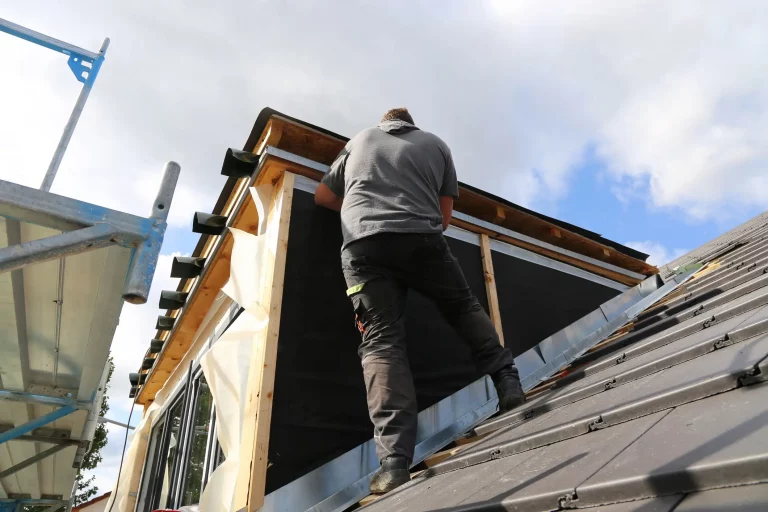 Ein Dachdecker in grauem T-Shirt und dunkler Arbeitskleidung steht auf einem geneigten Dach und arbeitet an dem Bau einer Dachgaube. Im Hintergrund ist ein Gerüst zu sehen, das zur Sicherheit und Unterstützung dient. Das Wetter ist teilweise bewölkt mit blauen Himmelabschnitten. Der Arbeiter verwendet Werkzeuge und Baumaterialien, um die Konstruktion zu sichern.