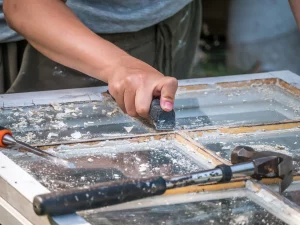 Ein Handwerker entfernt alte Farbe und Lack von einem Holzfensterrahmen mit einem Schaber. Auf dem Arbeitstisch sind Werkzeuge wie ein Hammer und ein Meißel für die Sanierung zu sehen.