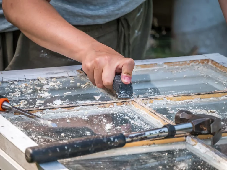 Ein Handwerker entfernt alte Farbe und Lack von einem Holzfensterrahmen mit einem Schaber. Auf dem Arbeitstisch sind Werkzeuge wie ein Hammer und ein Meißel für die Sanierung zu sehen.
