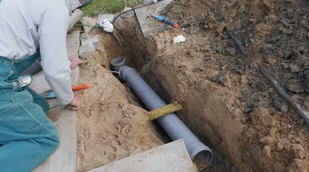Ein Handwerker in grüner Arbeitskleidung und grauem Sweatshirt führt eine Kanalsanierung durch. Er kniet neben einem offenen Graben im Außenbereich, in dem ein graues Abwasserrohr verlegt ist. Um den Graben herum liegen Werkzeuge und Erdhaufen.