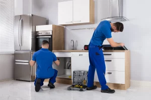 Zwei Küchenbauer in blauer Arbeitskleidung montieren eine neue Küche. Einer kniet vor einem offenen Unterschrank und arbeitet daran, während der andere sich über eine Arbeitsplatte beugt und ein Gerät installiert. Auf dem Boden steht ein offener Werkzeugkasten. Die Küche ist modern eingerichtet, mit einem großen Edelstahlkühlschrank und weißen Schränken.
